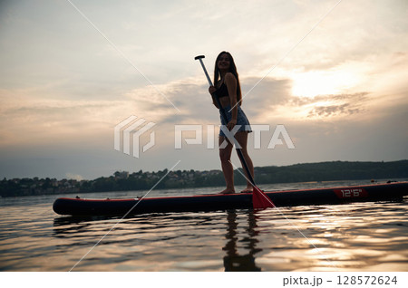 Against cloudy sky. Young woman is on sup boards in the lake 128572624