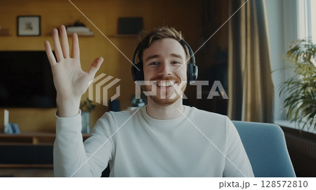 Smiling Young Man Greeting With a Wave While Wearing Headphones in a Cozy Living Room Setting 128572810