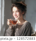 A beautiful young woman with a messy bun smiles contentedly, holding a glass cup of tea, as she looks out a window, bathed in warm, natural sunlight, evoking a peaceful morning moment 128572883