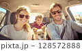 A joyful family, including parents, a child, and a small dog, all wearing sunglasses and smiling broadly from inside a sunlit car, capturing a happy road trip moment 128572888