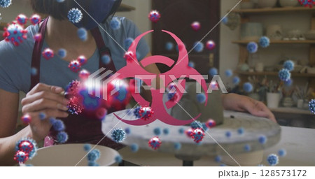 Image of covid 19 cells and biohazard sign over woman working in pottery studio in face mask Image of covid 19 cells and biohazard sign over woman working in pottery studio in face mask 128573172