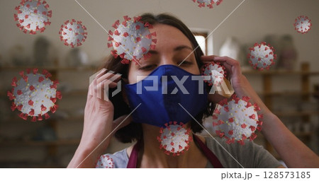 Image of covid 19 cells over woman working in pottery studio putting face mask on 128573185