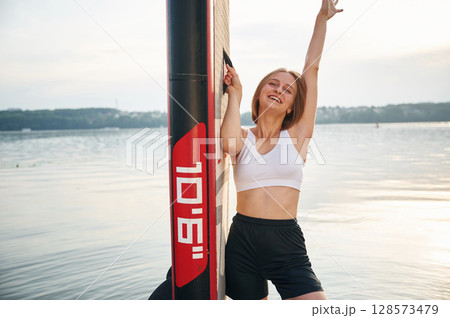 Smiling and enjoying the summer. Woman is standing with sup board against the lake Smiling and enjoying the summer. Woman is standing with sup board against the lake 128573479