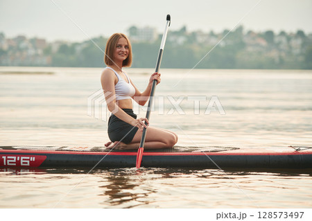 Forest and houses on background. Young woman is on sup boards in the lake Forest and houses on background. Young woman is on sup boards in the lake 128573497
