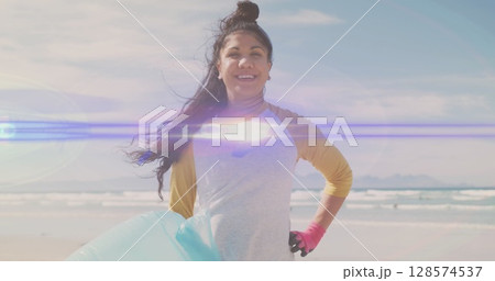Image of lights over smiling hispanic woman picking up rubbish from beach 128574537