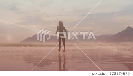 Image of light trails over caucasian woman walking with surfboard at beach 128574679
