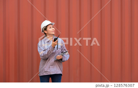 Female port worker in white helmet and uniform holding  walkie talkie and tablet for coordinating logistics loading operations at container terminal shipping yard 128576307