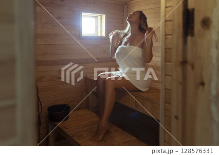 A young woman relaxes and sweats in a hot sauna wrapped in a towel. girl in the sauna. Finnish sauna interior, classic wooden sauna with hot steam. Russian bath. 128576331