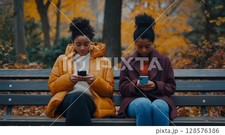 Two Friends Using Smartphones on a Park Bench During Autumn 128576386