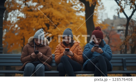 Friends Sitting on a Bench in a Park, Enjoying Smartphone Time Surrounded by Autumn Leaves 128576408