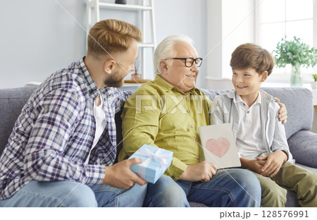 Elderly man with beaming smile receives gifts from his adult son and little grandson. 128576991