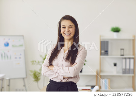 Young woman looking cheerful at camera standing with crossed arms in modern office. 128577256