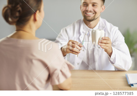 Dentist holding dental toy tooth with toothbrush in hands sitting in dentistry clinic with patient. 128577385
