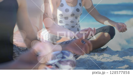 Image of light spots over diverse women practicing yoga and meditating at beach 128577858