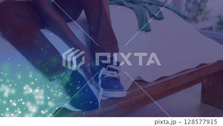 Image of lights over feet of african american boy tying shoe on bed 128577915