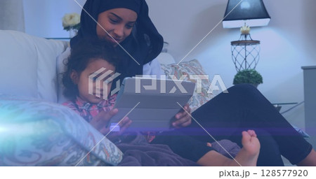 Image of glowing spots over happy biracial mother in hijab with daughter using tablet 128577920