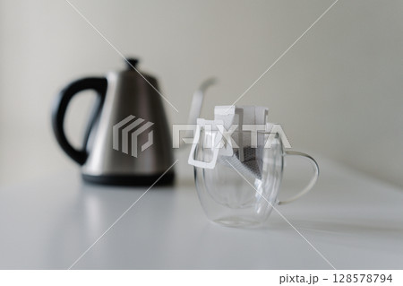 Drip coffee bag placed in glass cup, ready brewed with hot water from modern electric kettle placing on white background, close-up. Concept of convenient, quick way to enjoy fresh cup of coffee. 128578794