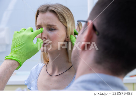 Young Woman Receiving Medical Consultation for Facial Treatment with Professional Practitioner in Health Clinic Setting, Focus on Patient's Expression and Doctor's Hand Preparation Young Woman Receiving Medical Consultation for Facial Treatment with Professional Practitioner in Health Clinic Setting, Focus on Patient's Expression and Doctor's Hand Preparation 128579488