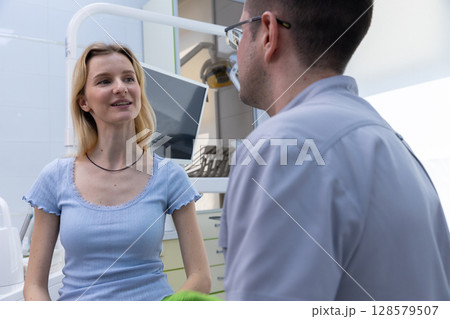 A Young Woman Engaging in Conversation with a Dentist in a Modern Dental Office Setting, Showcasing Professionalism and Patient Comfort During a Dental Visit A Young Woman Engaging in Conversation with a Dentist in a Modern Dental Office Setting, Showcasing Professionalism and Patient Comfort During a Dental Visit 128579507