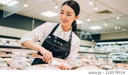 魚を手に取り品質を確認するスーパーの女性店員 128579514