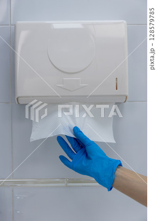 Hand Reaching for Paper Towel from Wall-Mounted Dispenser with Blue Gloves in Clean Restroom Hand Reaching for Paper Towel from Wall-Mounted Dispenser with Blue Gloves in Clean Restroom 128579785
