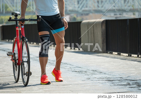 Cyclist walks with bandage on knee at public park. Unhappy cyclist with knee pain and injury outdoors. Cyclist walks with bandage on knee at public park. Unhappy cyclist with knee pain and injury outdoors. 128580012