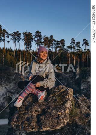 A woman in sportswear sitting on a rock at sunset, wearing a hat and looking like a hiker. The warm summer light and natural setting evoke themes of adventure, travel, and outdoor freedom. A woman in sportswear sitting on a rock at sunset, wearing a hat and looking like a hiker. The warm summer light and natural setting evoke themes of adventure, travel, and outdoor freedom. 128580306