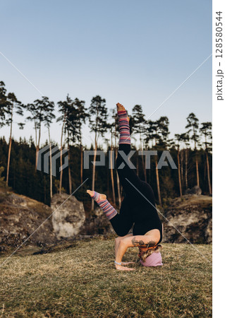 A woman practicing yoga at sunset in nature, barefoot and dressed in leg warmers and a hat,surrounded by rocks and forest. The scene reflects meditation, health, inner balance, and harmony with nature 128580544
