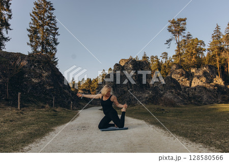 A woman practicing yoga at sunset in nature, barefoot and dressed in leg warmers and a hat,surrounded by rocks and forest. The scene reflects meditation, health, inner balance, and harmony with nature 128580566