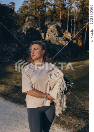 A woman in sportswear holding a yoga mat, standing among rocks and forest, ready for outdoor practice and mindfulness in nature 128580569