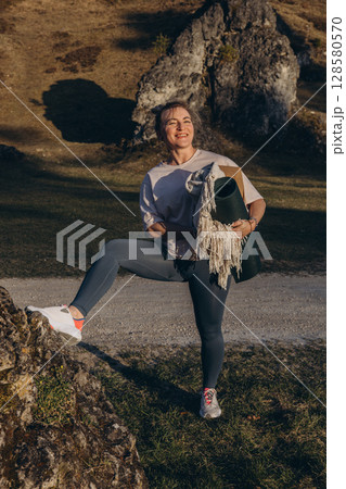 A woman in sportswear holding a yoga mat, standing among rocks and forest, ready for outdoor practice and mindfulness in nature A woman in sportswear holding a yoga mat, standing among rocks and forest, ready for outdoor practice and mindfulness in nature 128580570