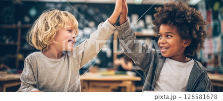 Two happy young boys in a classroom share a high-five, celebrating a moment of success or camaraderie, showcasing friendship and positive interaction in an educational setting 128581678