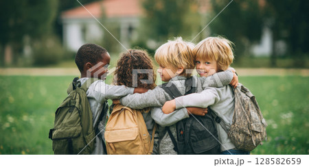 Four young children with backpacks, seen from behind with arms around each other, share a moment of friendship and togetherness in a green outdoor setting before school 128582659