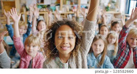 A happy young girl with curly hair confidently raises her hand and smiles at the camera, surrounded by a classroom full of other eager students also participating 128582660