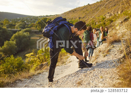 Man tourist tying his shoelace during walking tour in nature with people climbing a mountain Man tourist tying his shoelace during walking tour in nature with people climbing a mountain 128583712
