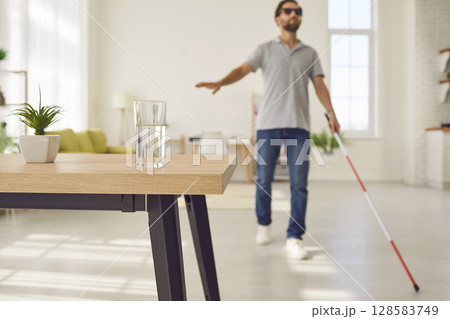 Blind man with stick is walking towards table with glass of water in living room 128583749