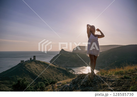 A blonde woman stands on a hill overlooking the ocean. She is wearing a white dress and she is enjoying the view. 128585730