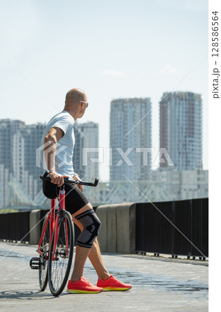 Cyclist pauses to enjoy city skyline on sunny day. Standing next to red bike, wearing athletic gear and sunglasses, he thoughtfully at modern buildings. 128586564