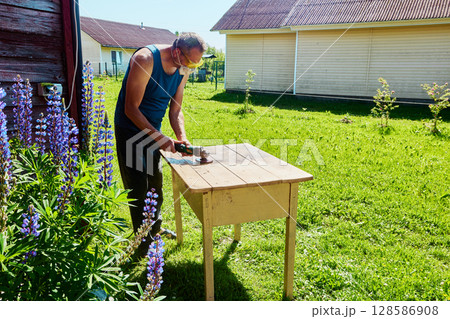 Sanding wooden tabletop with angle grinder outdoors, hobbyist working in yard near wooden houses on sunny summer day in countryside. 128586908