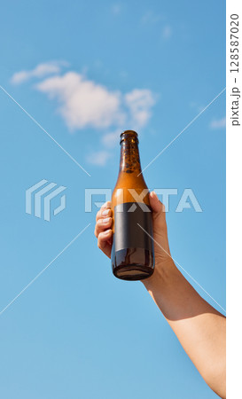 Hand holding beer bottle with blank label in front of clear blue sky with white clouds Hand holding beer bottle with blank label in front of clear blue sky with white clouds 128587020