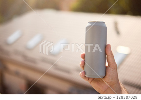 Grey matte drink can held in hand with blurred house rooftop in background 128587029