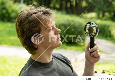Handsome young caucasian man with hand electric fan suffering from heat at park. Heat Summer. Handheld fan. Beat heat. 128587634