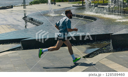 Midlife man jogging beside urban fountains on sunny day 128589460