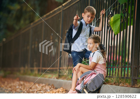 By the fence, relaxing. Young school children of boy and girl are together outdoors By the fence, relaxing. Young school children of boy and girl are together outdoors 128590622