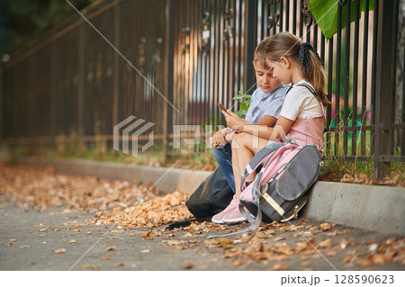 Sitting and using smartphone. Young school children of boy and girl are together outdoors Sitting and using smartphone. Young school children of boy and girl are together outdoors 128590623