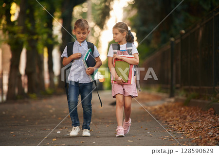 Front view, walking together. Young school children of boy and girl are outdoors Front view, walking together. Young school children of boy and girl are outdoors 128590629