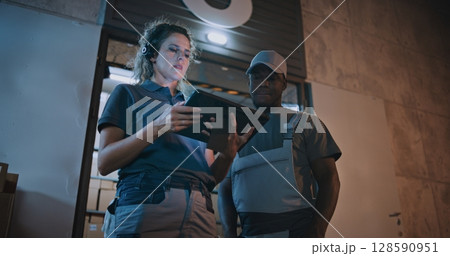 Employee and Female Manager Working in Delivery Service at Night: They Talking, Using Digital Tablet Employee and Female Manager Working in Delivery Service at Night: They Talking, Using Digital Tablet 128590951