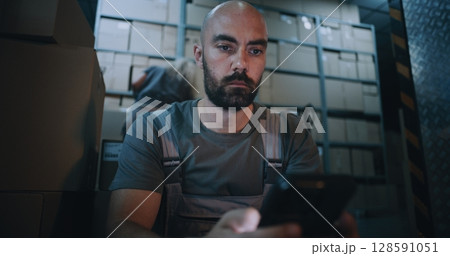 Exhausted Loader Using Smartphone, Having Rest after Hard Workday in Logistics Warehouse at Night 128591051