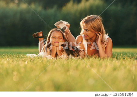 Lying down on the grass. Mother with her daughter and cute dog are on the field outdoors Lying down on the grass. Mother with her daughter and cute dog are on the field outdoors 128591294