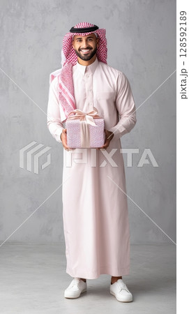 Smiling middle-eastern young male in traditional attire holding gift box. 128592189